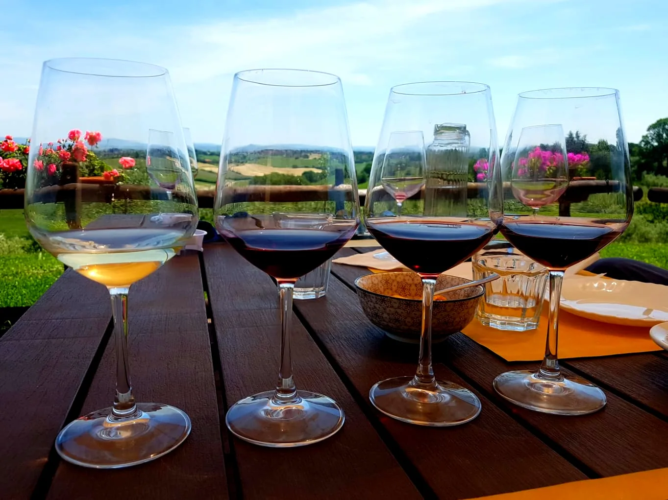 Quattro calici di vino su un tavolo all&rsquo;aperto, con le colline toscane sullo sfondo