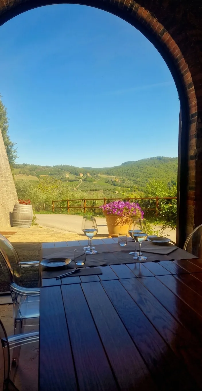 Tavolo apparecchiato con calici sotto un arco in mattoni, affacciato sulle colline toscane